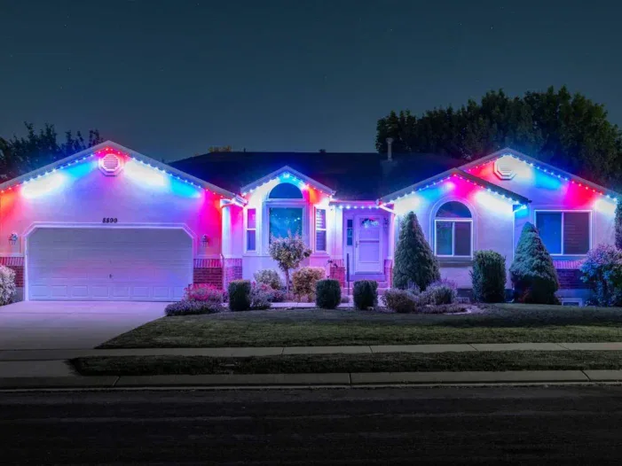 Single-story house at night with bushes in front, illuminated by bright red, white, and blue lights along the roof, windows, and entrance, creating a festive appearance. The driveway and lawn are visible under the lights.