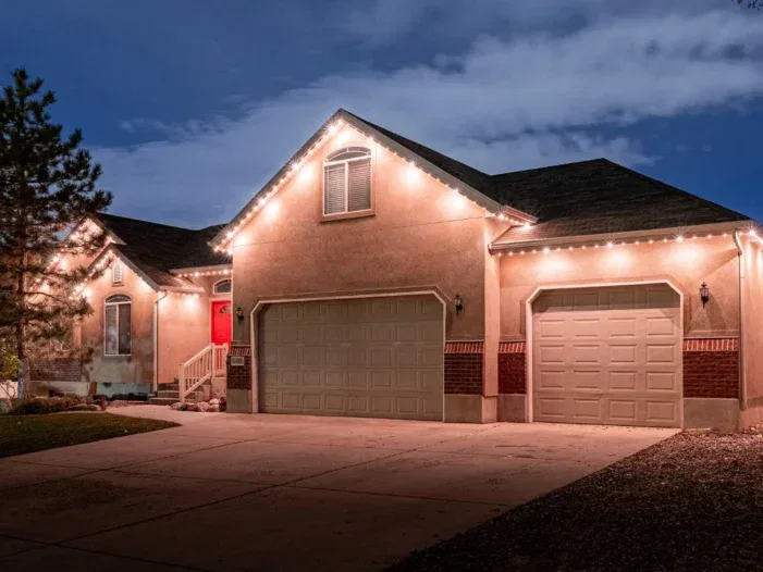 A single-story house with a red front door and three-car garage is lit by white string lights along the roofline at dusk. A tree is on the left, and the sky is partly cloudy.