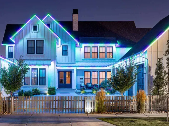 A modern two-story house at night, decorated with vibrant blue, green, and purple LED lights along the roofline. The home features large windows, a porch, a picket fence, and neatly landscaped yard with trees and shrubs.
