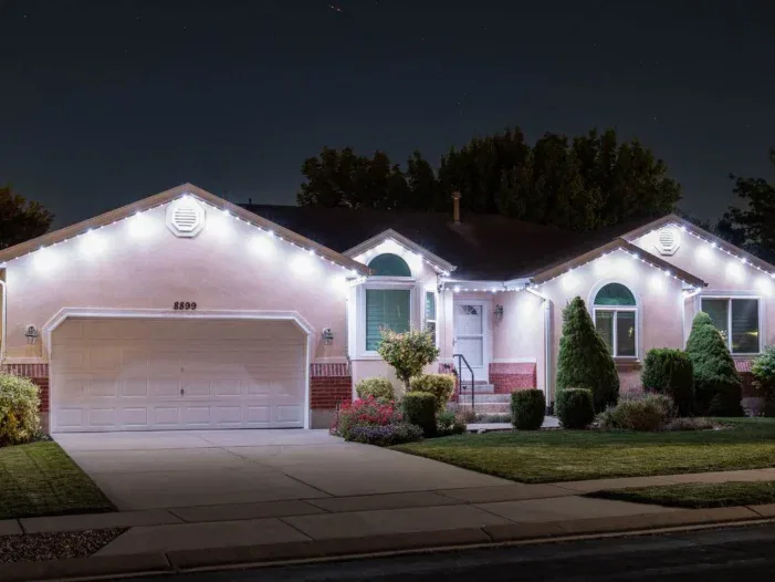 A single-story house at night with white trim and roofline Christmas lights, well-kept shrubs, a double garage, and a neatly manicured lawn.