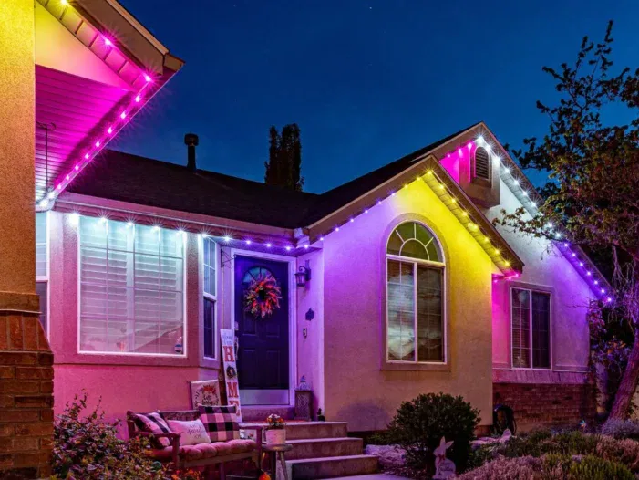 A single-story house at dusk is decorated with bright purple and white string lights along the roofline. A front porch with a wreath, bench, plants, and a bunny figurine is visible. The sky is deep blue.