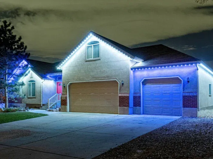 A single-story house with tan garage doors is decorated with bright white string lights along the roofline at night, illuminating the driveway and surrounding landscape.
