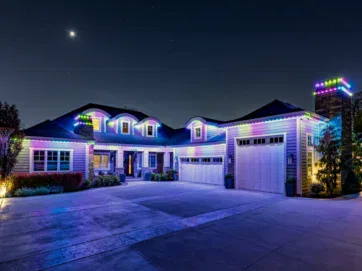 A large house at night is decorated with colorful LED lights outlining the roof, garage, and chimney. The driveway is empty and a crescent moon shines in the sky above.