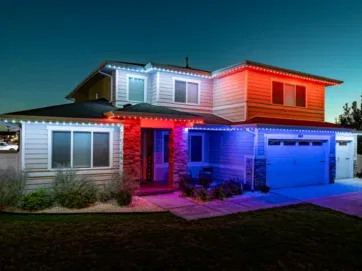 A two-story house at night with colorful lights outlining the roof, glowing red, white, and blue. The house has a garage, stone column accents, and a landscaped front yard. The sky is clear and dark.