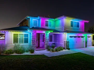 A two-story house at night, decorated with bright, colorful LED lights around the roofline, glowing in shades of blue, pink, green, and yellow. The yard and driveway are visible in the foreground.