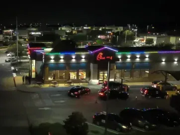 A Chick-fil-A restaurant at night is outlined with colorful lights. Cars line up in the drive-thru, and the parking lot is partially filled. City lights are visible in the background.