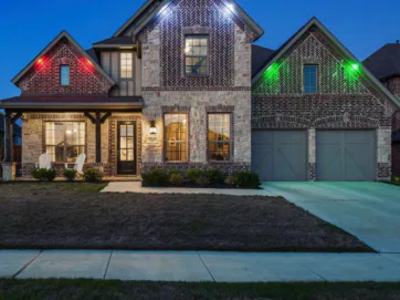A two-story brick and stone house with porch lights, a double garage, and colored lights (red, blue, green) illuminating the exterior, pictured in the evening with a dark blue sky.