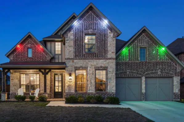 A two-story brick and stone house with porch lights, a double garage, and colored lights (red, blue, green) illuminating the exterior, pictured in the evening with a dark blue sky.