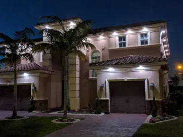 A two-story house with palm trees in front, illuminated by exterior lights at night, featuring two attached garages and a brick driveway.