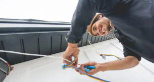 Attached image file A person smiling while installing or repairing electrical wiring on an exterior wall, holding colorful wires and using tools, viewed from a low angle.