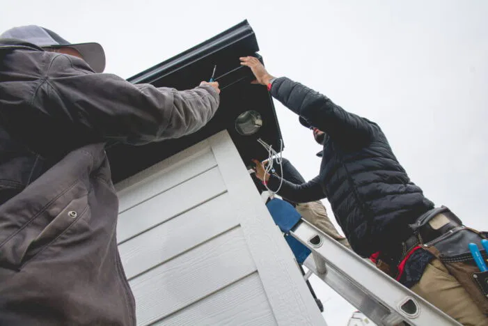 Two people on ladders install wiring under the roof of a small white building. One person wears a jacket and tool belt; both are using tools and working together on a cloudy day.