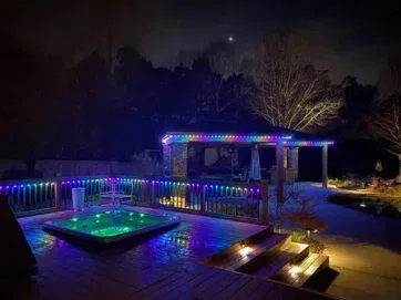 A backyard deck at night features a lit-up hot tub and gazebo, both outlined with colorful string lights. Trees and a dark sky are visible in the background, and soft lighting illuminates steps and pathways.