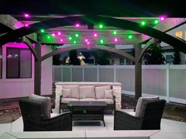A cozy outdoor seating area under a wooden pergola with green and purple string lights, featuring a cushioned sofa, two wicker chairs, and a fire pit table, surrounded by a white fence at dusk.
