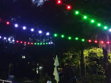 A nighttime outdoor scene with colorful string lights (red, green, purple) hanging above a swimming pool and tennis net, surrounded by trees and some poolside chairs.