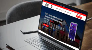 Attached image file A laptop on a table displays a website for Lee’s Summit Permanent Outdoor Holiday Lighting, featuring a house with colorful lights and a phone with a lighting control app. A closed notebook and pen are next to the laptop.