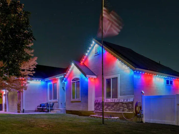 A single-story house at night is decorated with red, white, and blue lights. An American flag waves on a pole in the yard, and a bench sits near the front windows. The lawn and trees are softly illuminated.