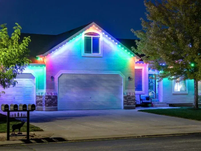 A suburban house at night is decorated with bright, colorful LED lights outlining the roof and garage, illuminating the driveway and trees. Four mailboxes are visible in front.