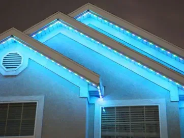 Exterior of a house at night with blue and white LED lights outlining the roof edges, creating a glowing effect against the dark sky. Two windows and a decorative vent are visible on the wall.