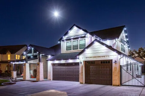 A modern two-story house with white siding and dark garage doors, decorated with white string lights. The driveway and front yard are illuminated under a bright night sky with a visible full moon.