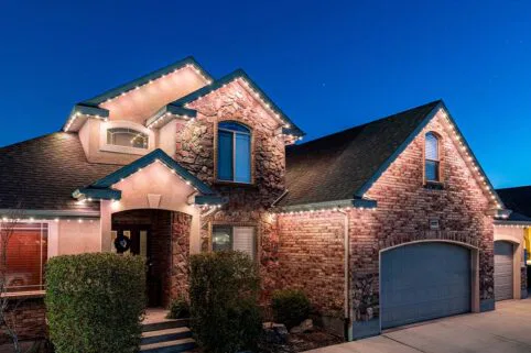 A two-story house with stone and brick exterior, lit by string lights along the roofline, with a closed garage door and trimmed bushes at the entrance, photographed at dusk under a clear sky.
