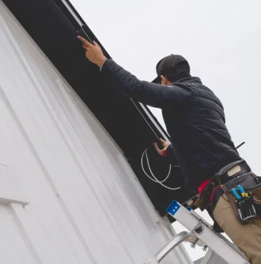A person standing on a ladder installs equipment or wiring near the roof of a white building, wearing a tool belt, jacket, and cap. The sky is overcast.