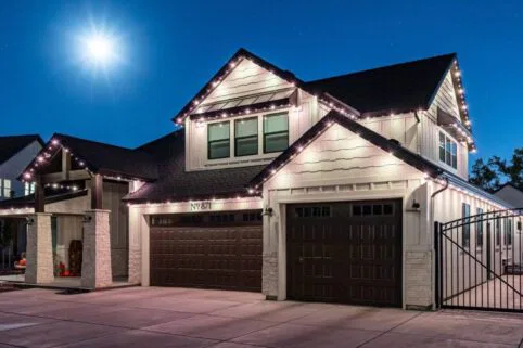 A two-story house with dark garage doors is decorated with white string lights on the roofline. The driveway and surroundings are lit by bright moonlight in a clear night sky.