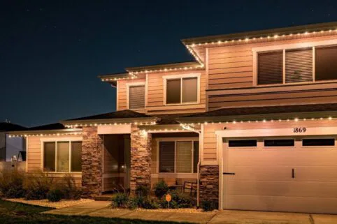 A two-story house with stone accents and tan siding is decorated with white string lights along the roofline. The house is lit up at night, with several windows and a visible house number: 1869.