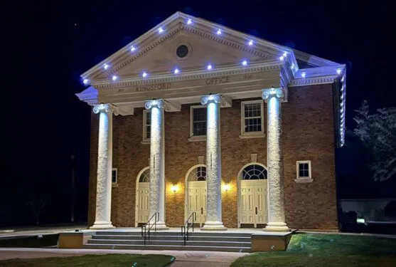 Attached image file A well-lit, two-story brick building with four large white columns and three arched doorways at night. White lights outline the roof, and Kingsford Office is engraved above the columns.