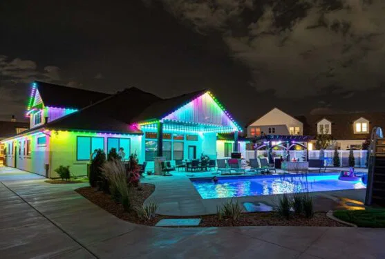 A modern house at night with multicolored LED lights outlining the roof, a well-lit swimming pool, lounge chairs, and a landscaped backyard under a partly cloudy sky.