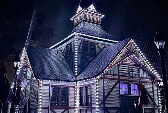 Attached image file Victorian-style building at night illuminated with white string lights along the rooflines and edges, highlighting its architectural details. The scene is dark, with glowing windows and light beams visible in the background.