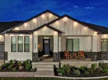 A modern single-story house at night, featuring white siding, dark stone accents, large windows, porch lights, two wooden chairs on the front porch, and a neatly landscaped front yard with small shrubs and plants.