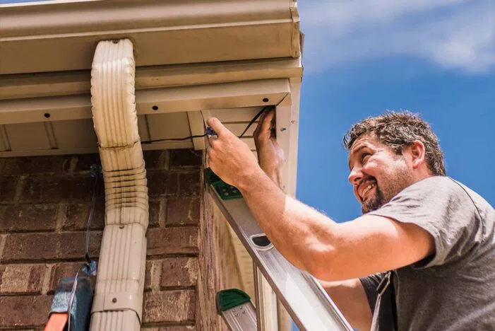 Attached image file A man stands on a ladder, repairing or inspecting a house gutter attached to a brick wall under a blue sky. He uses a tool and appears focused on his work.