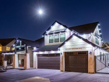 A two-story house with white walls and dark garage doors is decorated with white string lights along the roof. The driveway is empty, and a bright moon glows in the clear night sky above.