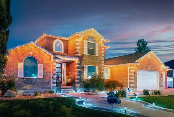A two-story brick house is decorated with string lights along the roofline and illuminated by garden lights at dusk. The yard features a manicured lawn, shrubs, rocks, and a curved walkway leading to the front entrance.