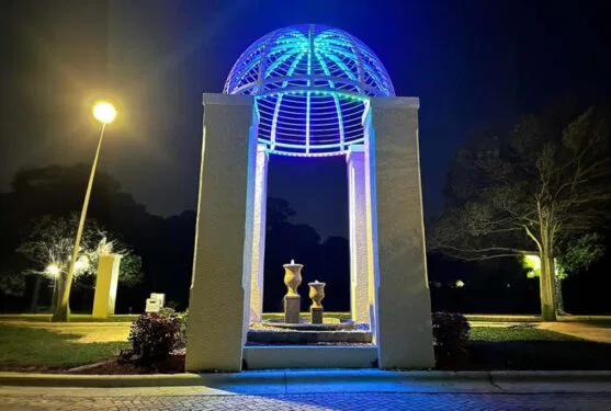 Attached image file A night scene of a small, open pavilion with a domed metal roof glowing blue light, two vases inside, and surrounding streetlights illuminating nearby trees and grass.