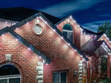 A two-story brick house at night is decorated with white string lights along the roofline, illuminating the exterior under a starry sky.