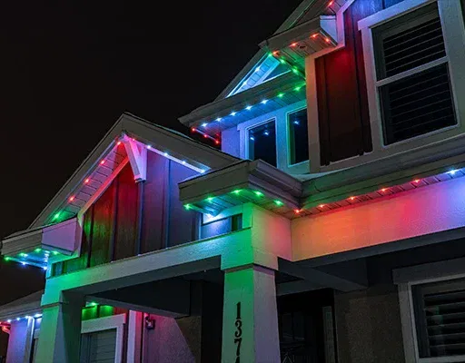 A house at night with colorful LED lights in red, blue, green, and white outlining the roof and eaves, creating a festive appearance. The house number 137 is visible on a white pillar.