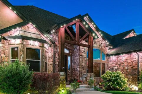 A stone house with large front windows and wooden beams is illuminated by exterior lights at dusk, with shrubs and flowers lining the entrance and a clear blue evening sky above.