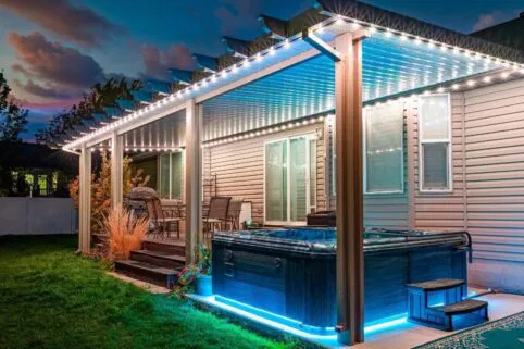 A backyard patio at dusk with string lights on a covered pergola, outdoor dining set on a wooden deck, and a hot tub illuminated with blue lights on a grassy lawn.