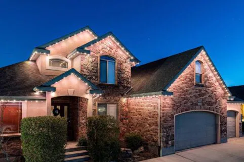 A two-story brick and stone house at dusk, with lit white string lights along the roofline, large windows, bushes near the entryway, and a gray garage door.