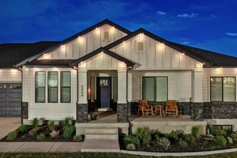 A modern, single-story house with white siding, dark stone accents, large windows, front porch with two wooden chairs and a small table, illuminated by outdoor lights at night. Shrubs line the walkway and entry steps.