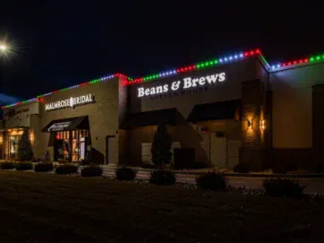 A coffee shop and bridal store with festive red, green, and blue lights on the roof, illuminated at night. The signs read “Beans & Brews Coffeehouse” and “Miamoré Bridal.”.