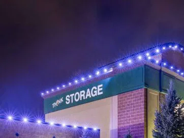 A building at night with THINK STORAGE written on the side, outlined with bright blue lights along the roof. Evergreen trees are visible in the foreground under a cloudy, purple sky.