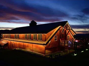 A large wooden barn illuminated by warm string lights at dusk, with a dramatic blue and purple sky in the background. The building stands out against the dark landscape.