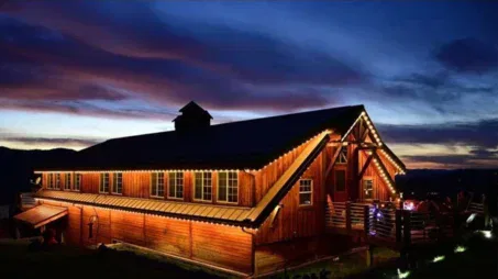 A large wooden barn illuminated by warm string lights at dusk, with a dramatic blue and purple sky in the background. The building stands out against the dark landscape.