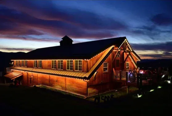 A large wooden barn illuminated by warm string lights at dusk, with a dramatic blue and purple sky in the background. The building stands out against the dark landscape.