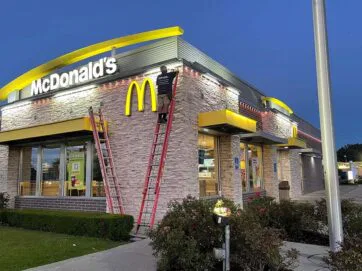 A person on a ladder works on the illuminated sign of a McDonald’s restaurant at dusk. The building features stone walls, yellow accents, and is surrounded by bushes and a parking lot.