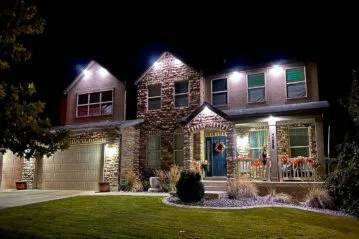 A two-story house with stone and siding exterior is lit up at night. The house has a front porch decorated with autumn wreaths and garlands, a blue front door, and a neatly manicured lawn.