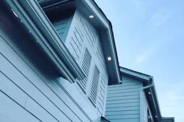 Close-up view of the upper exterior of a light blue house with horizontal siding, dormer windows with shutters, decorative shingles, and recessed lights under the eaves, against a clear blue sky.