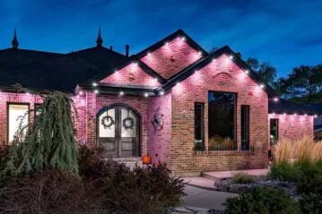 A brick house at dusk with glowing pink and white uplighting along the roofline, arched double doors, two wreaths, tall grasses, shrubs, and a small pumpkin on the porch. The sky is deep blue.